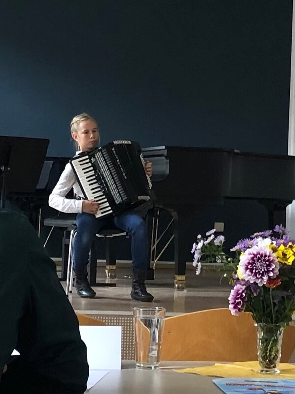 Eine Akkordeonschülerin sitzt auf einem Stuhl auf einer Tribune und spielt vor einer Jury.