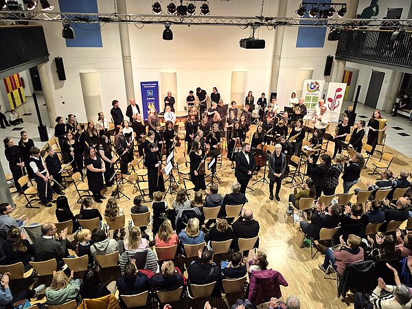 Man sieht ein Orchester. Alle Mitglieder stehen, während das Publikum applaudiert.