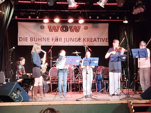 Auf einer Bühne sitzt und steht eine größere Gruppe junger Musikerinnen und Musiker in zwei Reihen. Sie spielen verschiedene Instrumente, darunter Saxophone, E‑Gitarre, Bass, Keyboard, Posaune und Schlagzeug. 