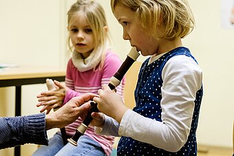 Ein Mädchen versucht mit der Hilfe der Lehrerin Blockflöäte zu spielen. Ein anderes Mädchen schaut zu.