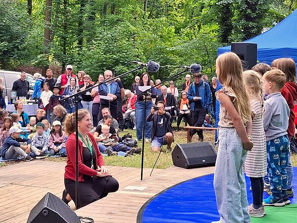 Ganz junge Musikschüler stehen auf der Bühne im Wald mit Mikrofonen und singen unter Leitung von Frau Linke vor einem aufmerksames Publikum.