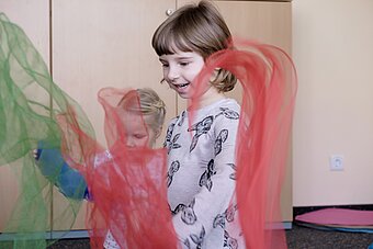 Es sind zwei Kinder aus der Musikalischen Früherziehung mit schwingenden Tüchern, in rot und grün,  zu sehen.