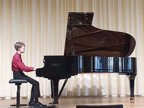 Auf dem Foto sitzt ein junger Pianist in einem roten Hemd und schwarzen Hosen an einem geöffneten Flügel. Er spielt konzentriert, die Hände über den Tasten, während beige Vorhänge den Hintergrund bilden.
