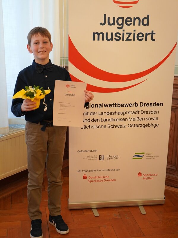 Ein Schüler steht vor dem Banner von Jugend musiziert und strahlt in die Kamera. Er hat seine Urkunde in der einen und einen Blumentopf in der anderen Hand.