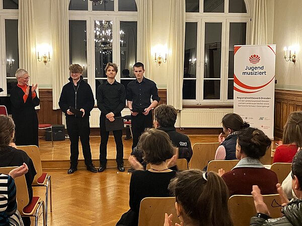 Drei Jugendliche stehen vorn in einem festlichen Raum und halten Urkunden. Sie werden von der Musikschulleiterin Frau Haas und dem Publikum mit Applaus geehrt. Rechts ist ein Banner mit dem Schriftzug „Jugend musiziert – Regionalwettbewerb Dresden“.