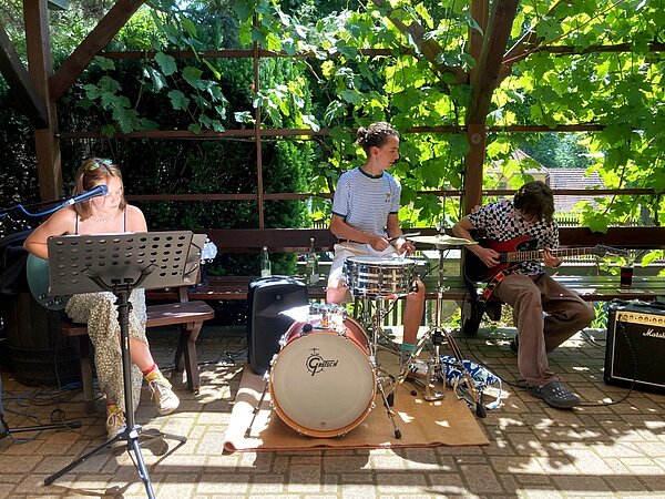 Drei Musiker spielen unter einer von grünen Weinranken überdachten Pergola im Freien. Links singt jemand mit Gitarre, in der Mitte sitzt ein Schlagzeuger, rechts spielt ein E-Gitarrist.