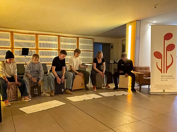 Ein Persussionensemble spielt in einem Foyer. Die Instrumentalisten sitzen auf Cajons. Rechts im Bild sieht man ein Banner stehen, welches das Musikschullogo zeigt.