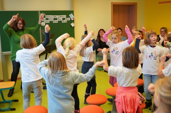 Die Kinder des Kinderchores  bewegen sich zur Musik. Alle schauen zur Chorleherin, diese steht im Hintergrund des Bildes. Sie macht die Bewegung vor.