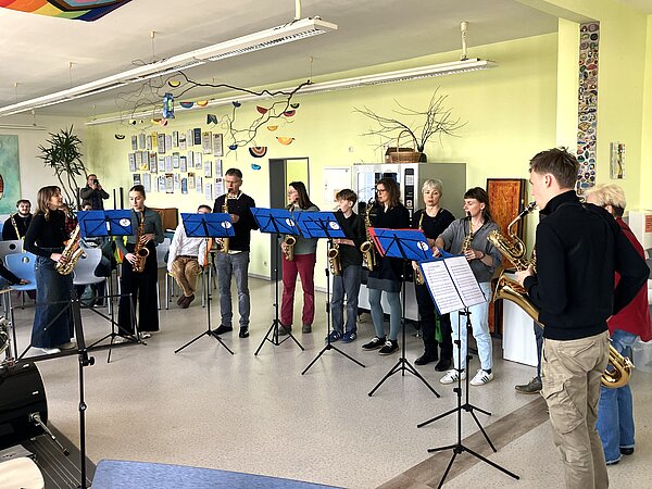 Auf dem Foto steht eine Gruppe von Saxophonspielerinnen und -spielern in einem Halbkreis in einem hellen, freundlichen Probenraum. Jede Person hält ein Saxophon, vor ihnen stehen mehrere Notenständer mit blauen Mappen.