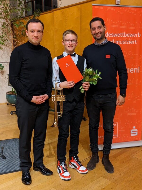 Unser Schüler Max steht in der Mitte, rechts steht sein Lehrer Herr Maka und links Herr Michalski. Max hält seine Trompete, eine Urkunde vom Landeswettbewerb "Jugend musiziert" und Blumen in den Händen.