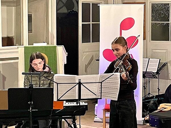 Es spielt eine Klavier- und eine Geigenschülerin als Duo. Im Hintergrund steht das Musikschulbanner und man sieht eine Kanzel.