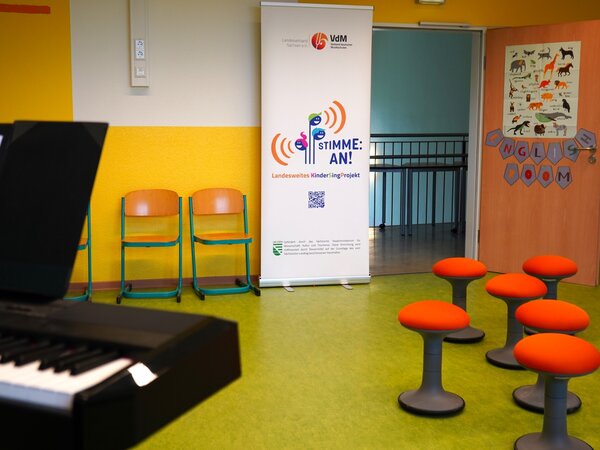 Es ist in der Mitte neben der geöffneten Zimmertür das Banner von Stimme an zu sehen. Stühle stehen auf anderen Seite an der Wand. Links sieht man die Ecke eines E-Pianos rechts stehen Hocker mit orangenem Sitzkissen.
