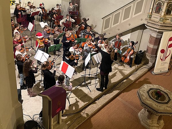 Es ist das Kinderstreichorchester zu sehen. Die Kinder stehen mit ihren Instrumenten im Altar der Friedenskirche und musizieren. 