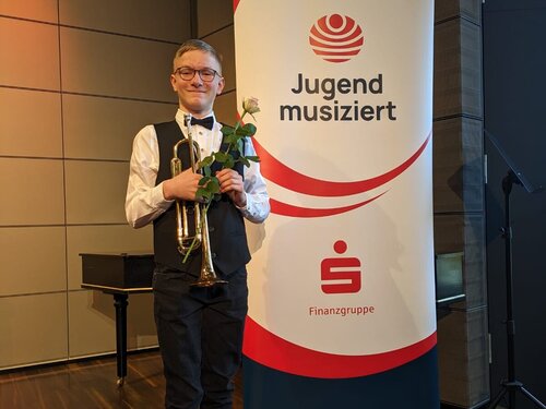 Ein Schüler steht vor dem Banner von "Jugend musiziert" und präsentiert sich freudig mit einer Rose und seiner Trompete. Die Rose erhielt er beim Landeswettberbausscheid "Jugend musiziert".