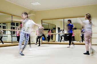 Es sind Mädchen in einem Tanzraum mit großen Spiegeln beim Tanzen zu sehen. 