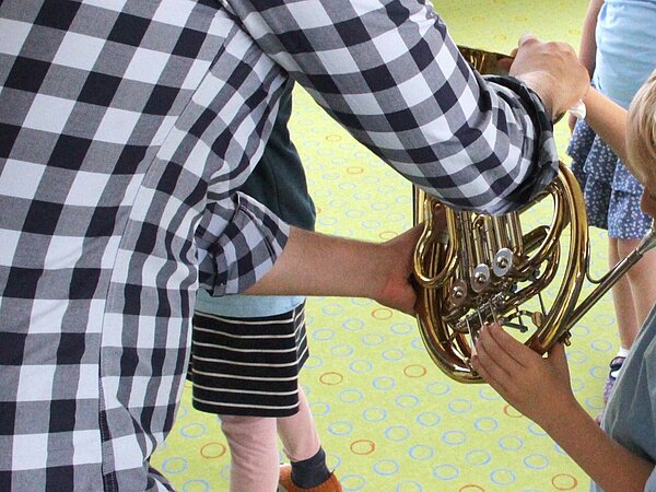 Man sieht wie ein Horn zum ausprobieren übergeben wird. Andere Kinder stehen dabei und schauen interessiert zu.