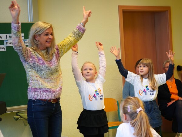 Uta Bresan steht mit Kindern in einem Klassenzimmer und halten die Hände in die Luft.