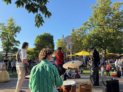 Die Band steht auf der Bühne und musiziert. Das Foto wurde mit Blick auf das Publikum aufgenommen und man sieht einen Hof voller Menschen und Kinder, welche sich vor der Bühne versammelt haben um der Musik zu lauschen.
