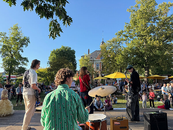 Die Band steht auf der Bühne und musiziert. Das Foto wurde mit Blick auf das Publikum aufgenommen und man sieht einen Hof voller Menschen und Kinder, welche sich vor der Bühne versammelt haben um der Musik zu lauschen.