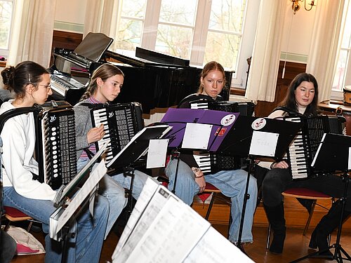 Vier Personen sitzen in einem Musikraum im Halbkreis und spielen Akkordeon. Vor ihnen stehen Notenständer, hinter ihnen ein Flügel und große Fenster, durch die Tageslicht fällt. 