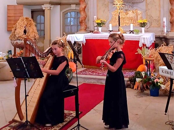 Zwei Musikschülerinnen sind vor dem Altar, eine Schülerin mit einer Harfe und die andere mit einer Geige. Sie musizieren gemeinsam.