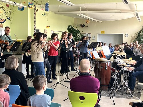 Auf dem Foto sieht man eine Gruppe von Musikerinnen und Musikern, die in einem freundlich gestalteten Raum ein kleines Konzert geben. Verschiedene Instrumente sind vertreten: Flöten, Saxophone, eine Geige, Congas, ein Drumset und weitere Percussion. Vor den Spielenden stehen Notenständer. Im Vordergrund sitzt ein kleines Publikum auf bunten Stühlen und hört aufmerksam zu. 