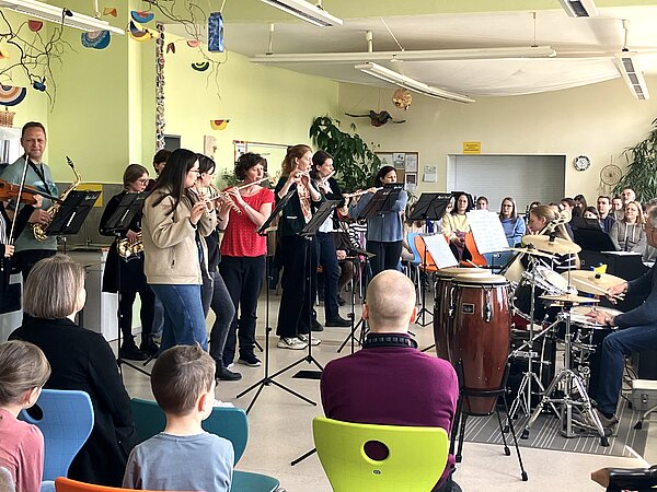 Auf dem Foto sieht man eine Gruppe von Musikerinnen und Musikern, die in einem freundlich gestalteten Raum ein kleines Konzert geben. Verschiedene Instrumente sind vertreten: Flöten, Saxophone, eine Geige, Congas, ein Drumset und weitere Percussion. Vor den Spielenden stehen Notenständer. Im Vordergrund sitzt ein kleines Publikum auf bunten Stühlen und hört aufmerksam zu. 