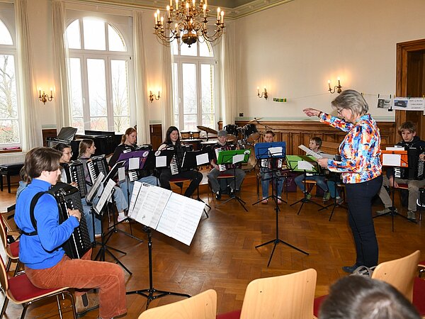 In einem hellen Musikraum sitzen mehrere Menschen in einem Halbkreis und spielen Akkordeon. Vor ihnen stehen Notenständer, im Hintergrund sind ein Klavier und ein Schlagzeug zu sehen. Eine Person vorne leitet die Gruppe an und gibt Anweisungen.
