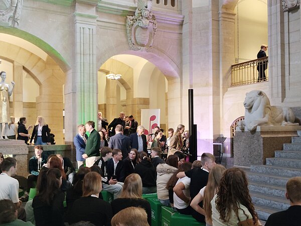 Eine Gesamtaufnahme in der sächsischen Staatskanzlei, mit mehreren Schülerinnen und Schülern, welche vor der großen Treppe sitzen und im Hintergrund schon das Musikschullogo zu sehen ist, sowie einige Rathausmitarbeiterinnen und Mitarbeiter.