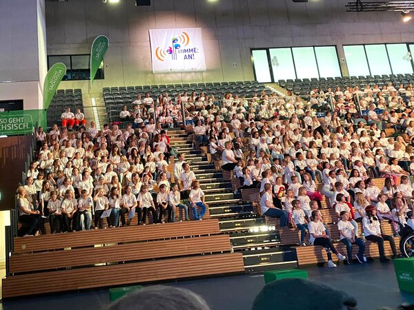 Es sind sehr viele Kinder auf der Tribüne der Ballsportarena sitzend zusehen. Sie nehmen am Projekt "Stimme an" teil.