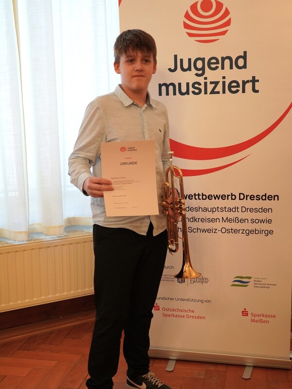 Ein Schüler steht vor dem Banner von Jugend musiziert und strahlt in die Kamera. Er hat seine Urkunde in der einen und eine Trompete in der anderen Hand.