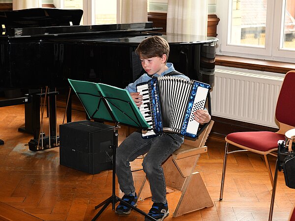 Ein junger Akkordeonschüler spielt auf einer Bühne. Dahinter sieht man einen Flügel stehen.