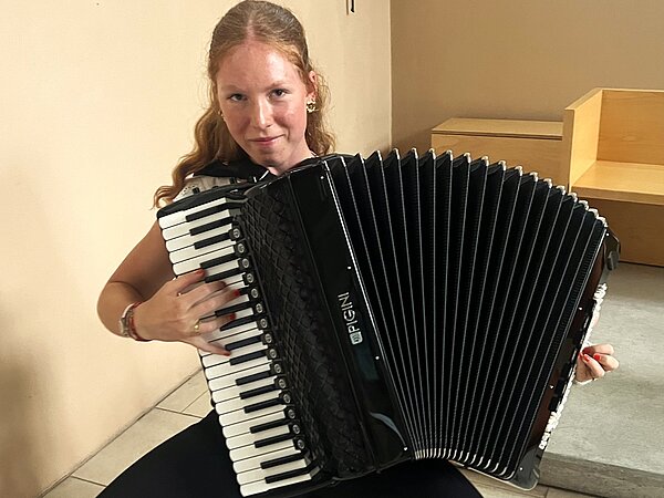 Eine Person sitzt drinnen, schaut in die Kamera und spielt ein großes schwarzes Akkordeon. Im Hintergrund stehen helle Holzmöbel und eine beige Wand.