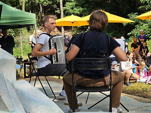 Zwei Akkordeonschüler spielen auf einer Bühne vor einem Publikum. Im Hintergrund sieht man den Wald.