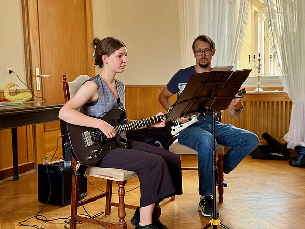 Eine E-Gitarrenspielerin sitzt auf einem Stuhl vor ihr ein Notenpult. Daneben sitzt ihr E-Gitarrenlehrer. Sie spielen zusammen auf ihren E-Gitarren.