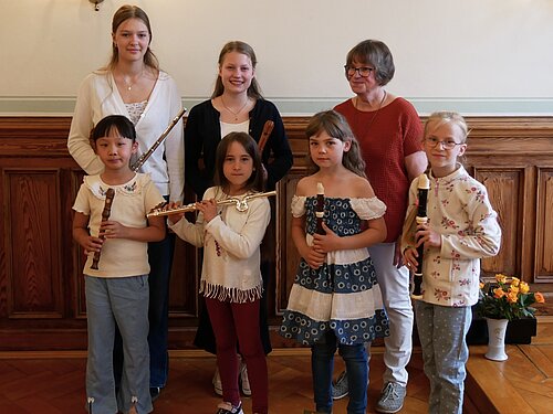 Sechs Flötenspierlerinnen mit ihren Flöten und ihre Lehrerin Frau Steinert lachen gemeinsam in die Kamera.