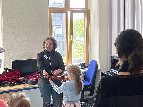 Ein Kind probiert mit Hilfe der einer Lehrkraft die Geige aus. Andere schauen dabei zu.