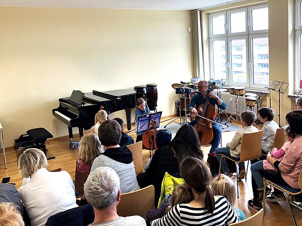 Das Foto zeigt eine Vorspielsituation in einem hellen Raum der Musikschule. Im Vordergrund spielen ein erwachsener Cellist und ein junges Mädchen gemeinsam auf ihren Instrumenten. Vor ihnen sitzt ein interessiertes Publikum aus Kindern und Erwachsenen.